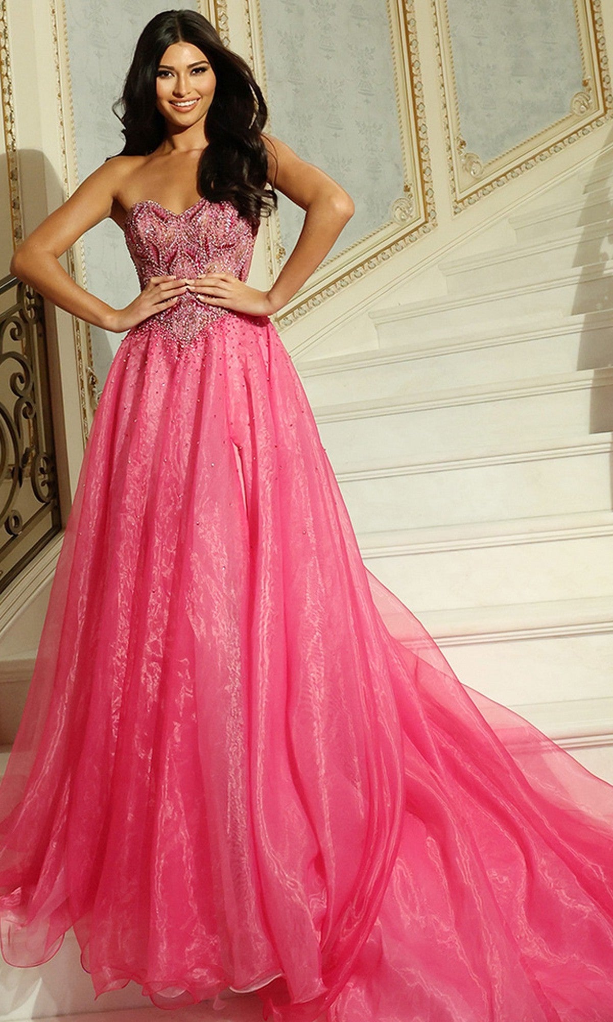 A woman poses on a staircase with ornate railings and decorative wall panels, wearing the Jovani 48906 long prom dress featuring a strapless beaded corset bodice and an a-line organza skirt.